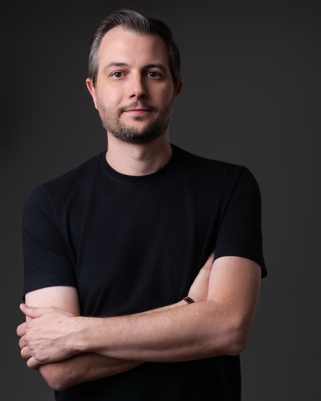 Retrato de Daniel Sagin, de camiseta preta, braços cruzados, sobre fundo cinza